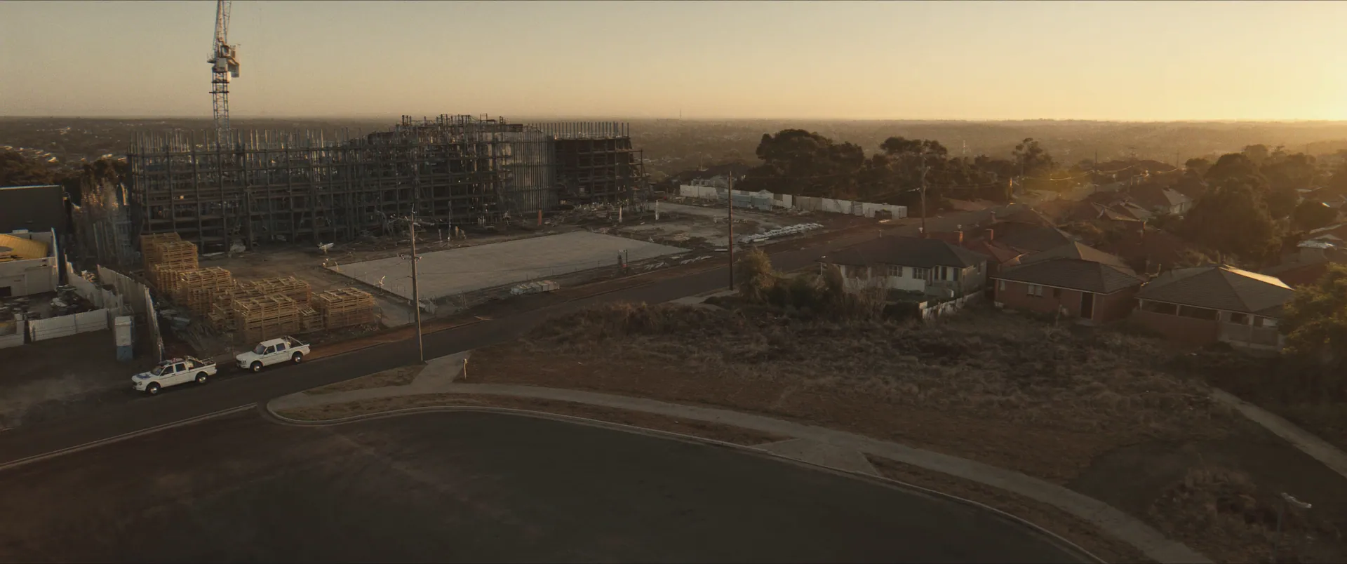 Aerial of a suburban construction site at golden hour with a tower crane and parked utes